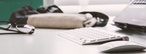 White desk with keyboard, mouse, glasses and a bag on it.