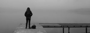 Female standing on a doc looking out with a backpack next to her (black & white).