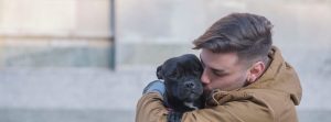Man holding and kissing a small black dog outside.