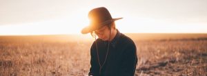 Man with glasses and a hat looking down standing outside with he sun behind him.