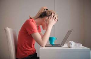woman in pink shirt holding head stressed by laptop and teal mug.