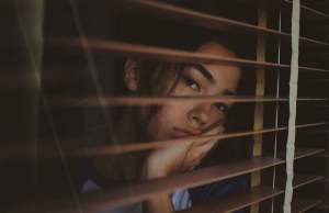 Woman looking out window with wooden blinds.
