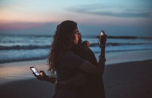 two people hugging and holding cell phones at sunset on the beach.