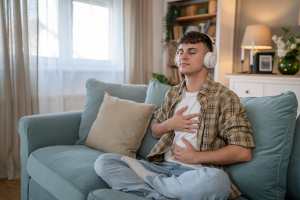 Man sitting on a couch wearing headphones and pressing his hands on his chest during meditation
