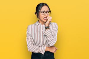 Woman with glasses biting her nails anxiously in front of a yellow background.