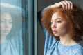 Red-haired woman looking out the window deep in thought, her reflection visible in the window.