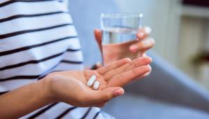 Hand cupping two pills with a glass of water in the other hand while the person is sitting on a couch