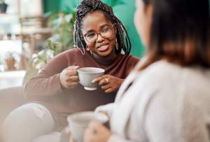 Woman in a red sweater drinking tea on a couch with her friend.